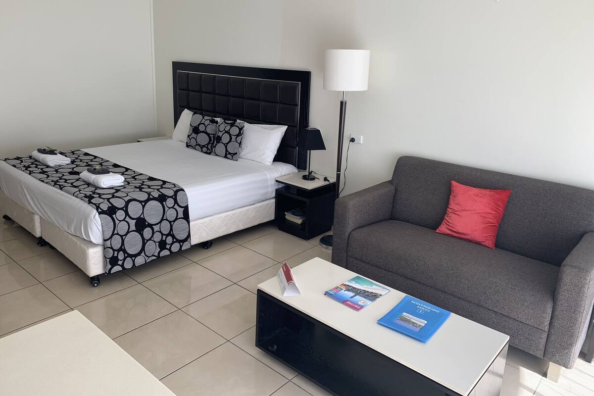A hotel room with a bed, a grey sofa, a coffee table with books, a nightstand with a lamp, and a tiled floor.