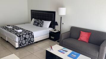 Een hotelkamer met een bed, een grijze bank, een salontafel met boeken, een nachtkastje met een lamp en een betegelde vloer.