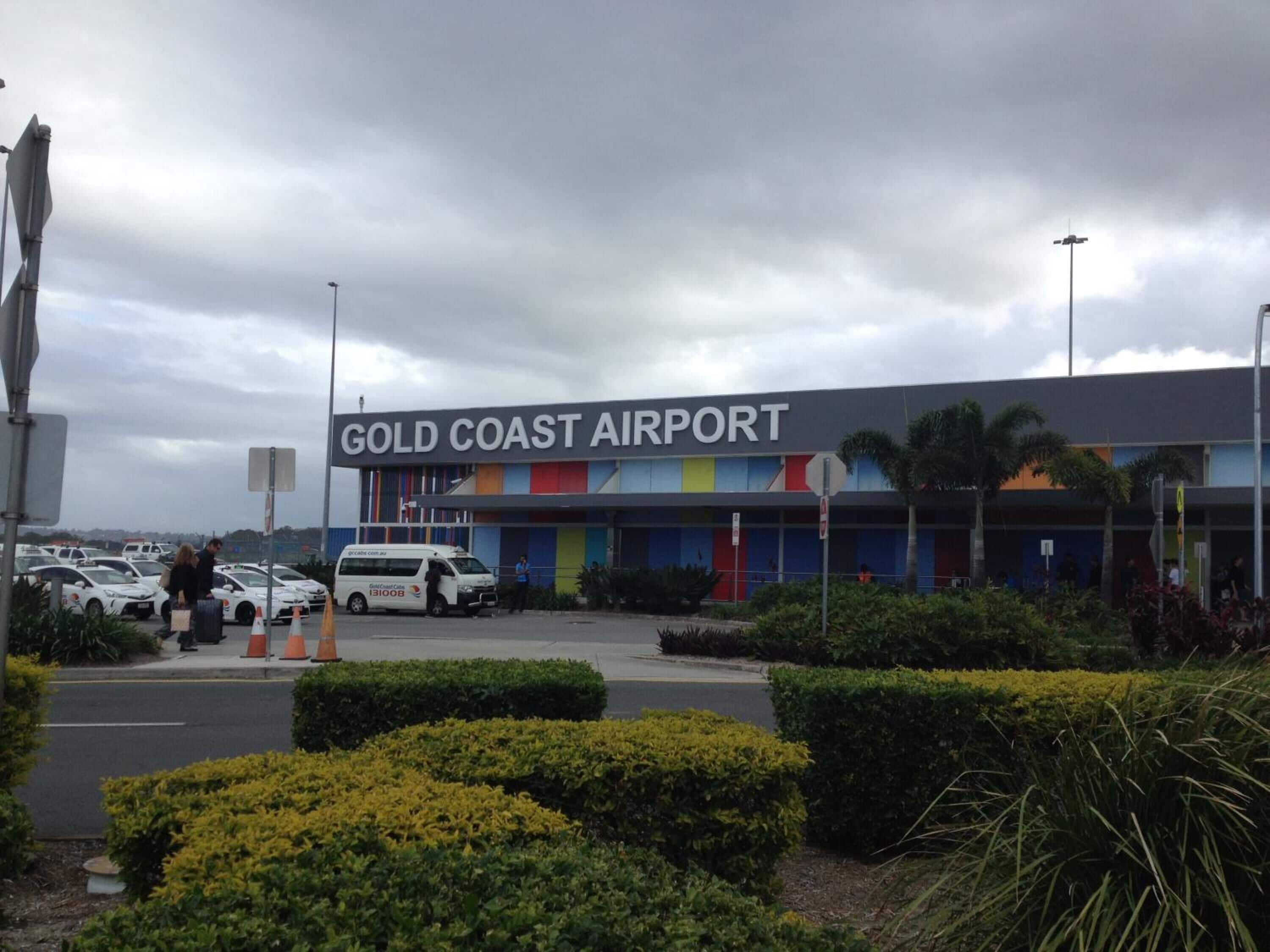 The exterior of Gold Coast Airport with a modern design, featuring a large sign and a parking area with vehicles.