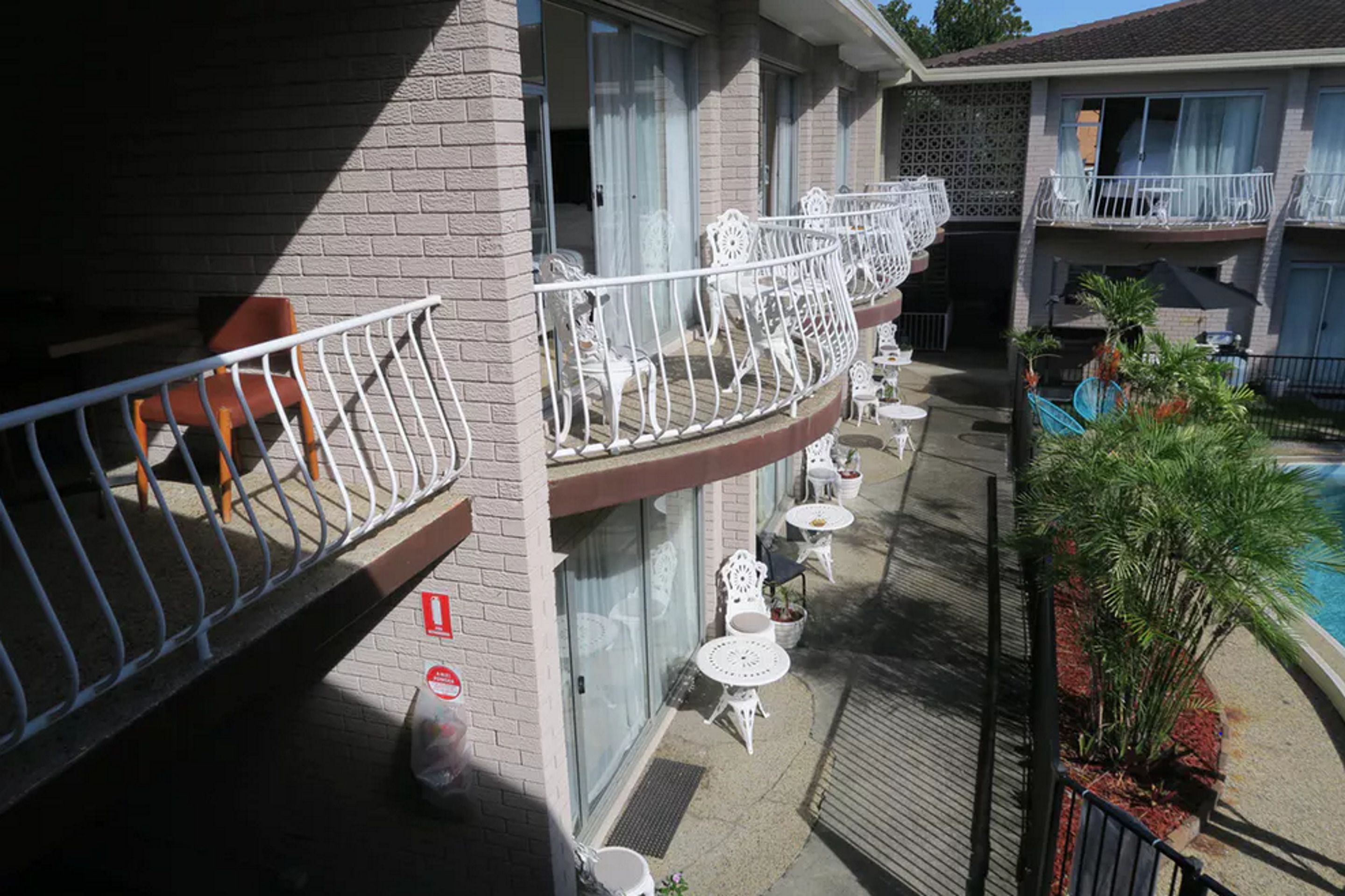 A hotel with a balcony with white chairs and tables, a pool area with palm trees, and a brick facade.