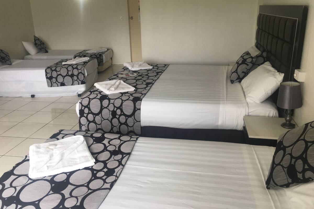 A hotel room with two beds, each with a patterned bedspread, a bedside table with a lamp, and a door in the background.