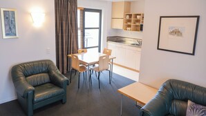 A compact living space with a dining area, two leather armchairs, a small table, a kitchenette, and a large window with curtains. - Victoria Court Motor Lodge (Wellington)