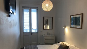 Un dormitorio con cama, mesita de noche, televisor montado en la pared, ventana con persianas, lámpara colgante, espejo y un cuadro enmarcado.