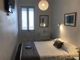 A bedroom with a bed, a bedside table, a TV mounted on the wall, a window with blinds, a hanging light fixture, a mirror, and a framed picture on the wall.