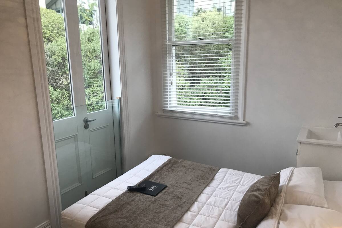 A bedroom with a bed, a window with blinds, a door, and a bedside table with a lamp.