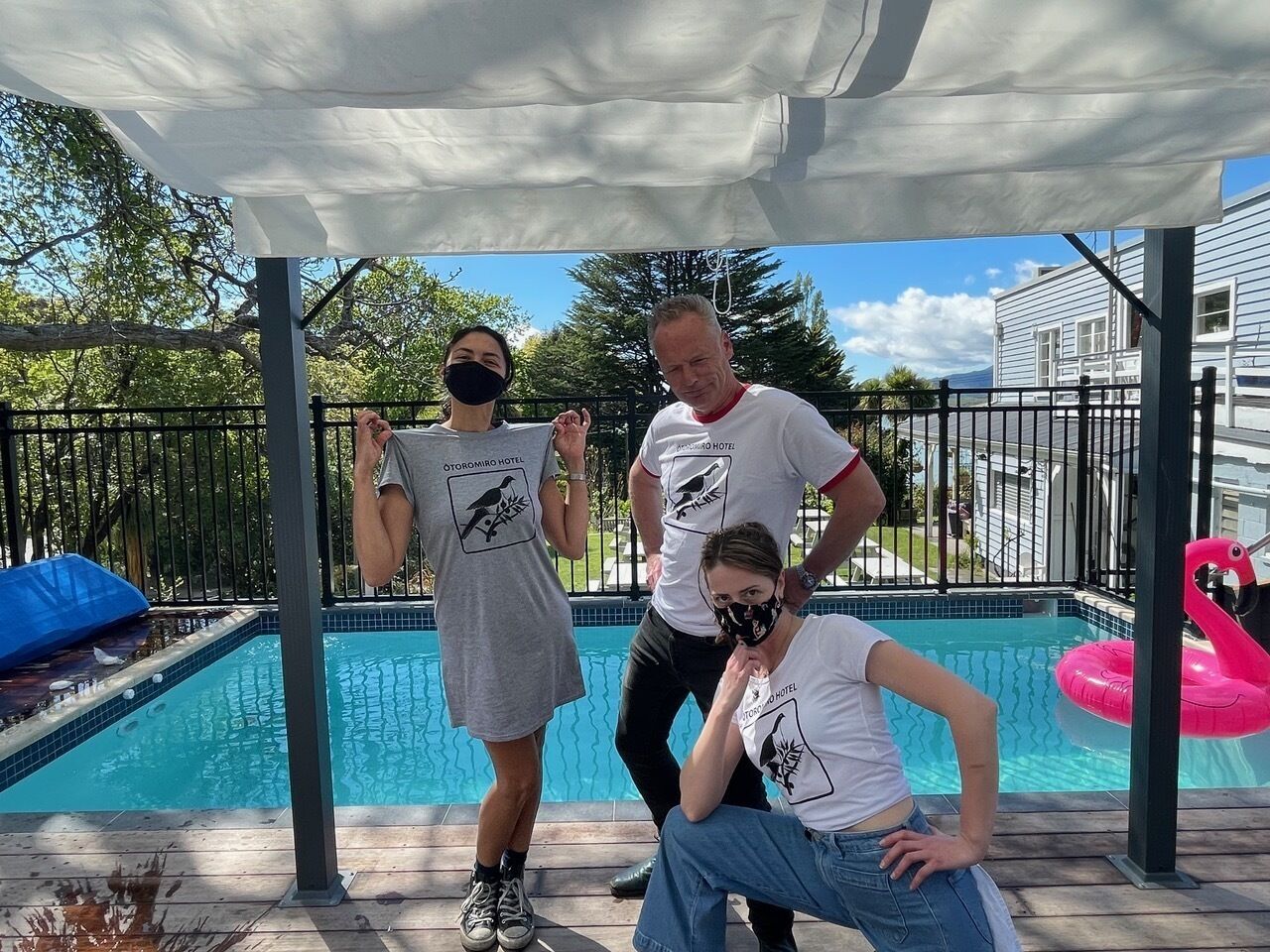 Un espace piscine doté d’un coin salon couvert, d’un flotteur en forme de flamant rose et de motifs d’oiseaux imprimés sur les t-shirts des personnes présentes.