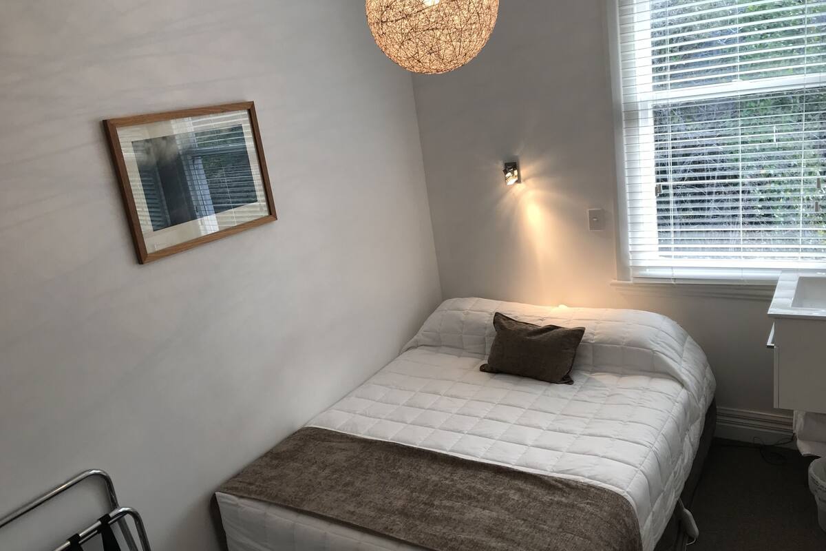 A neatly arranged bedroom with a bed, a bedside table, a hanging light fixture, a window with blinds, and a framed picture on the wall.