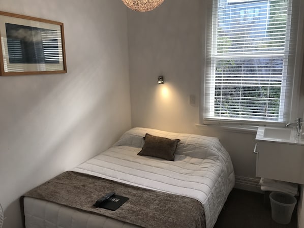 Un dormitorio pequeño y ordenado con una cama individual, una mesita de noche, una lámpara de pared y un cuadro en la pared. Cuenta también con una ventana con persianas.