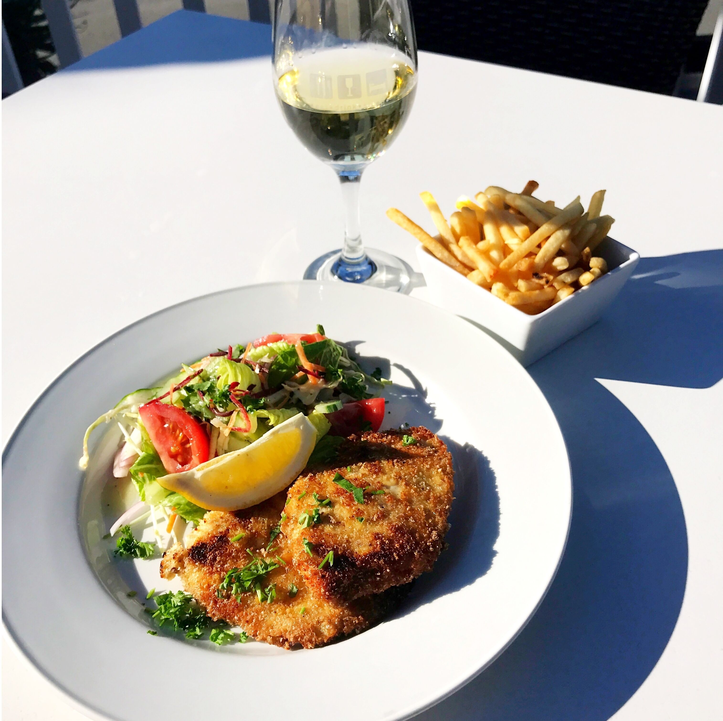 Un plato con un alimento empanizado y frito, una ensalada de tomates y lechuga, y una copa de vino blanco sobre una mesa blanca.
