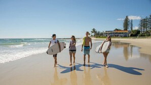 A beach scene with four people walking along the shore, each carrying a surfboard. - YHA Coolangatta Gold Coast (Bilinga)