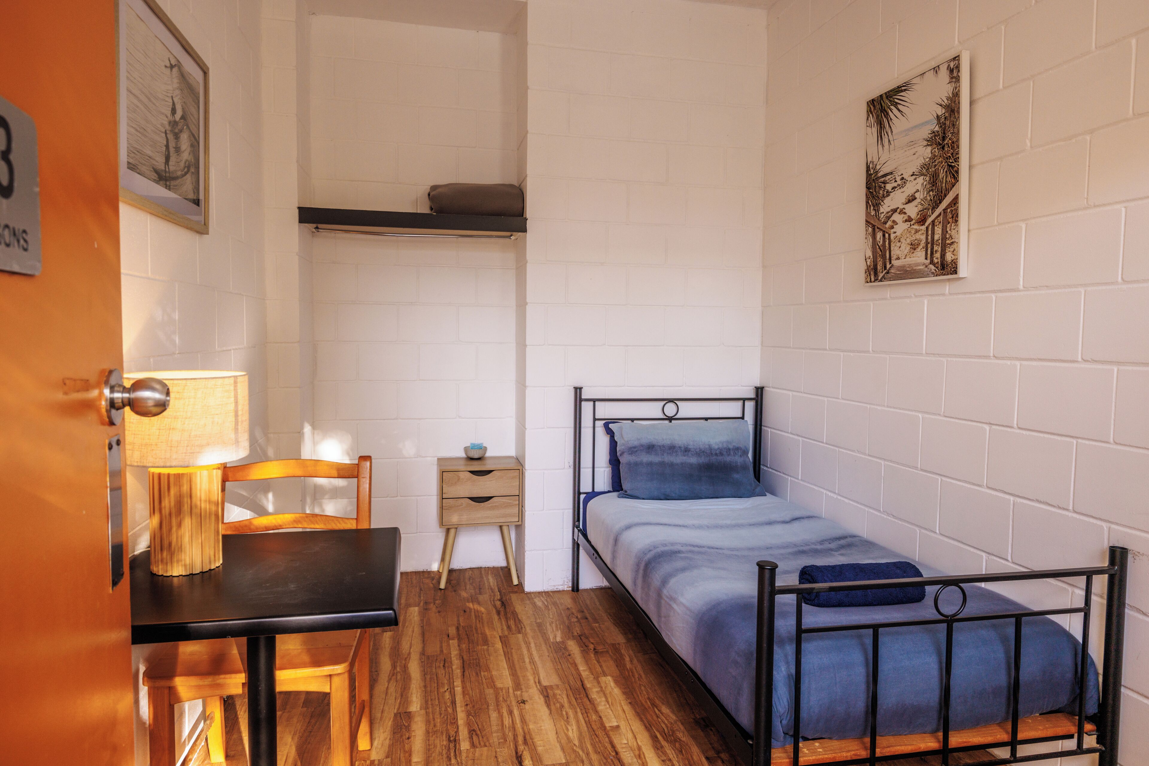 Una habitación pequeña y funcional, con cama, una mesa de madera, una silla, una cómoda pequeña y un estante en la pared.