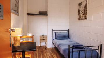 Una habitación pequeña y funcional, con cama, una mesa de madera, una silla, una cómoda pequeña y un estante en la pared.