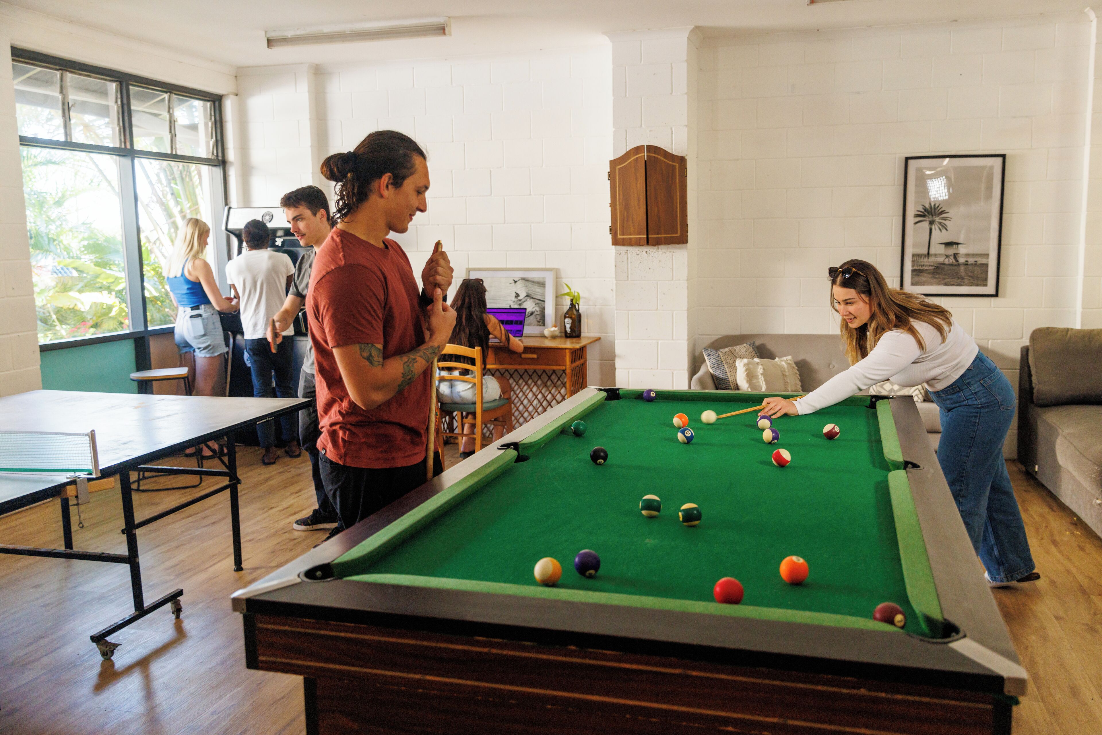 A pool table in a room with white brick walls, a wooden floor, and a window with a view of greenery.