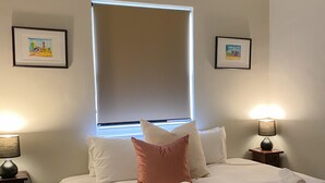 Un lit bien fait, avec du linge de lit blanc et un coussin rose, deux tables de chevet avec des lampes et des tableaux encadrés accrochés aux murs.
