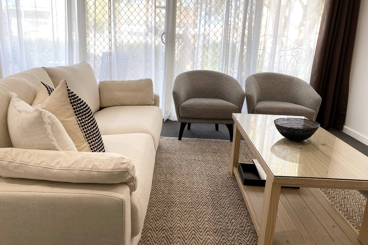 A living room with a beige sofa, two armchairs, a glass coffee table, and a patterned rug.