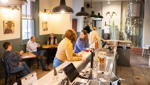 Zona de bar con clientes sentados en mesas, una barra con grifo y una gran instalación de elaboración industrial en el fondo.
