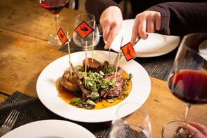 A dining table with a plate of food, glasses of red wine, and a person reaching for a glass.