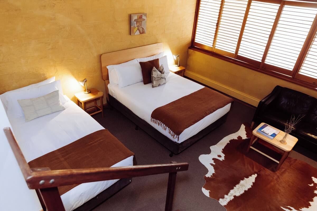 A hotel room with two beds, a wooden headboard, a nightstand with a lamp, a leather chair, and a cowhide rug.