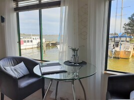 Ein Zimmer mit Glastisch, zwei Stühlen und Blick auf Boote auf dem Wasser, sowie ein Fenster mit Vorhängen.