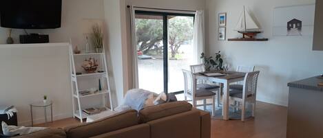 A modern living room with a sofa, a dining table with chairs, a TV mounted on the wall, and a bookshelf.