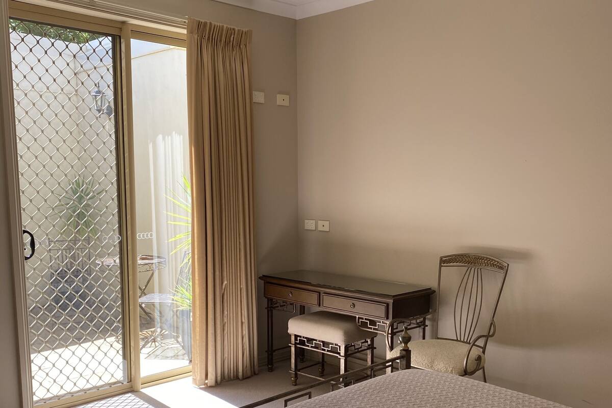 A bedroom with a large bed, a desk, and a chair. There is a sliding glass door leading to a balcony.