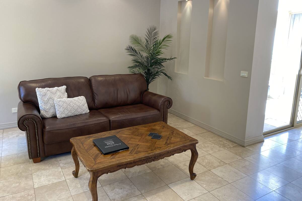 A brown leather sofa with two white cushions, a wooden coffee table with a book, and a large plant in a living room with tiled flooring and recessed lighting.