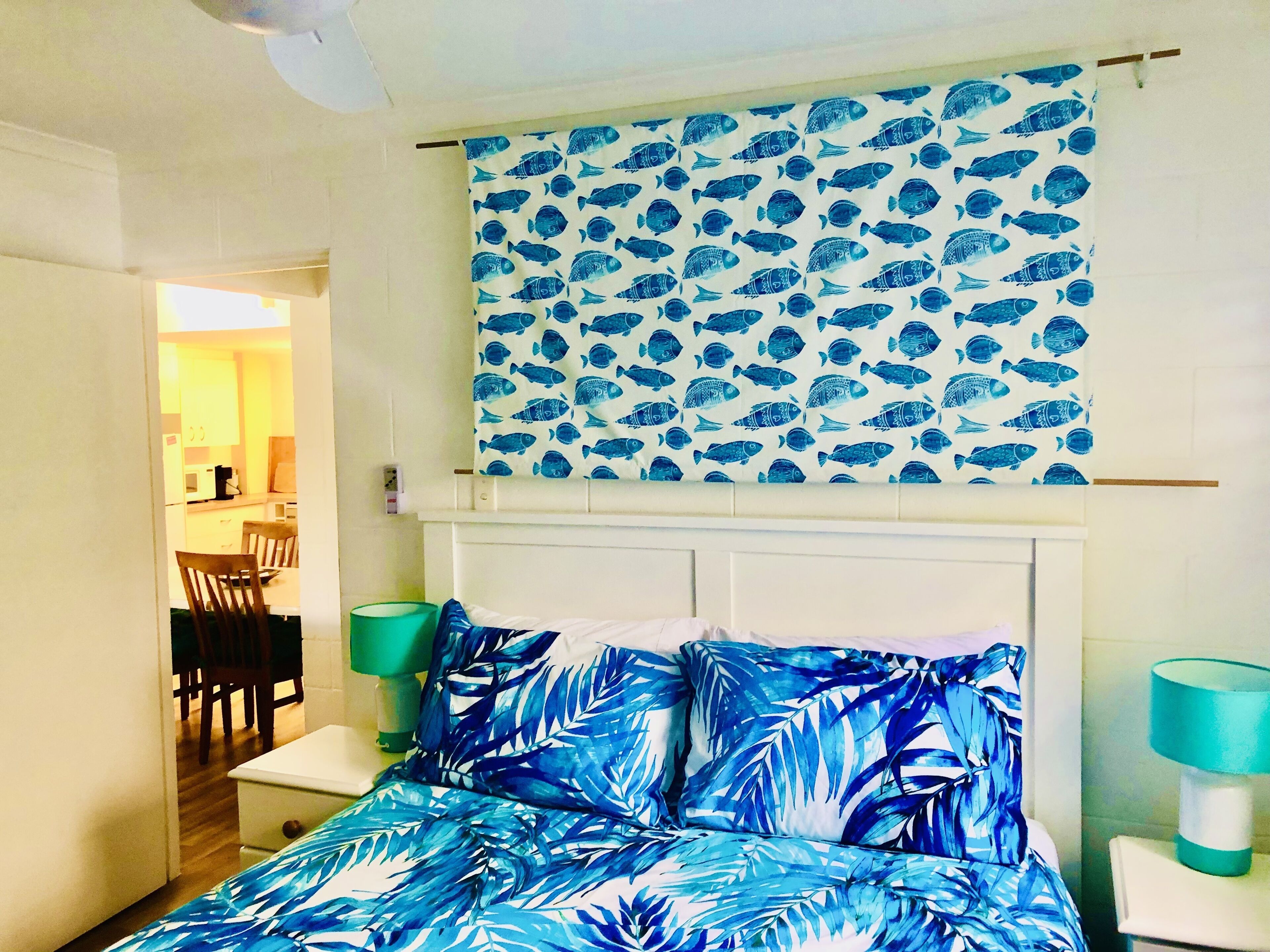 Un dormitorio con cama, cabecera, ventana con cortinas, mesita de noche y una silla.