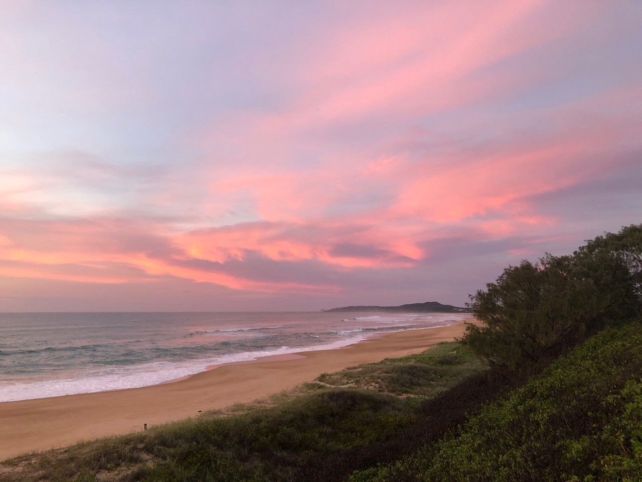 A beach with a view of the ocean and a distant landmass under a pink and purple sky.