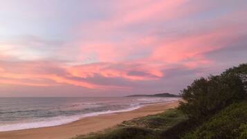 A beach with a view of the ocean and a distant landmass under a pink and purple sky.
