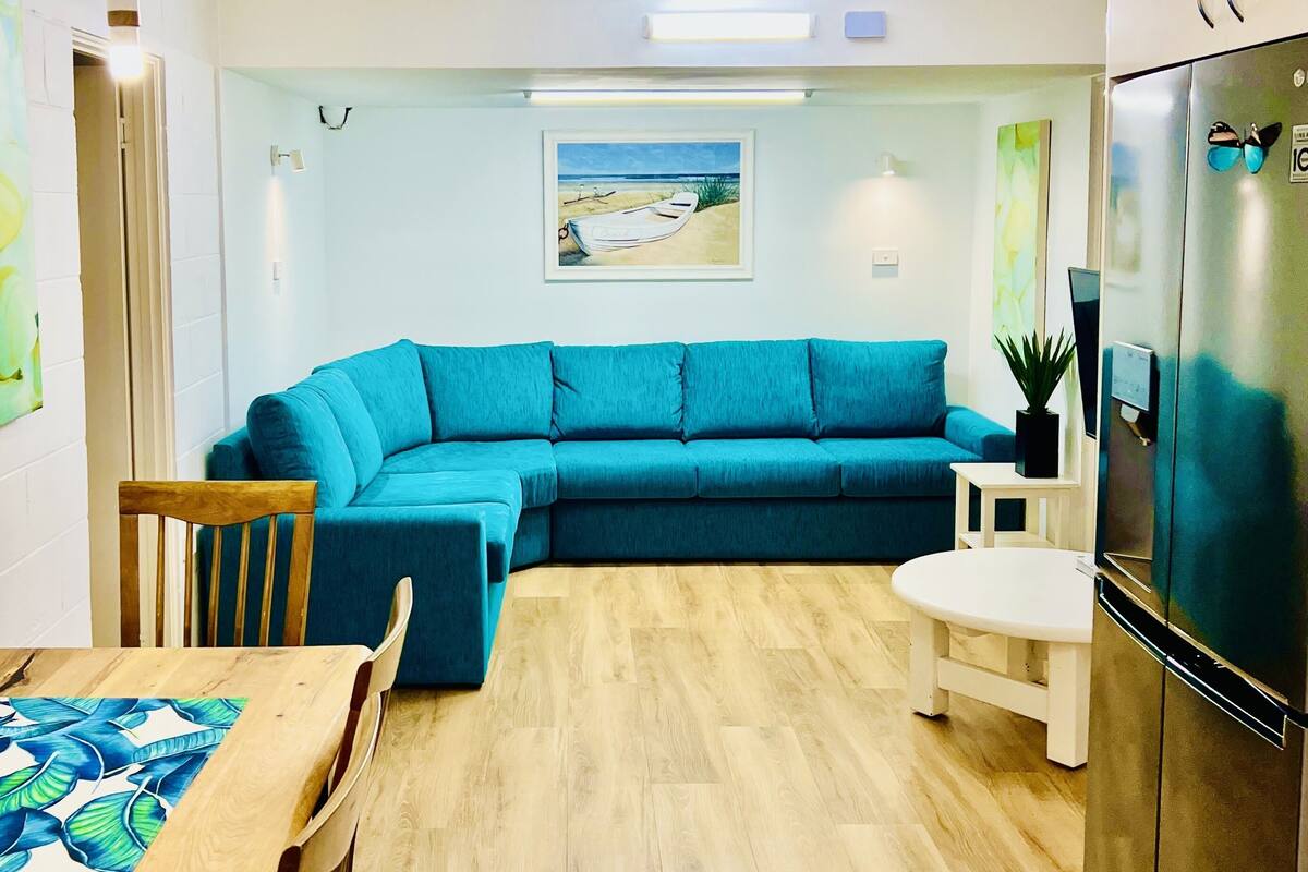 A modern living room with a teal sofa, a wooden dining table, and a kitchen area with a refrigerator.