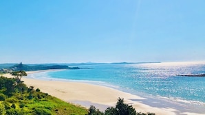 Ein Strand mit klarem, blauem Wasser, sandigem Ufer und Vegetation im Vordergrund.