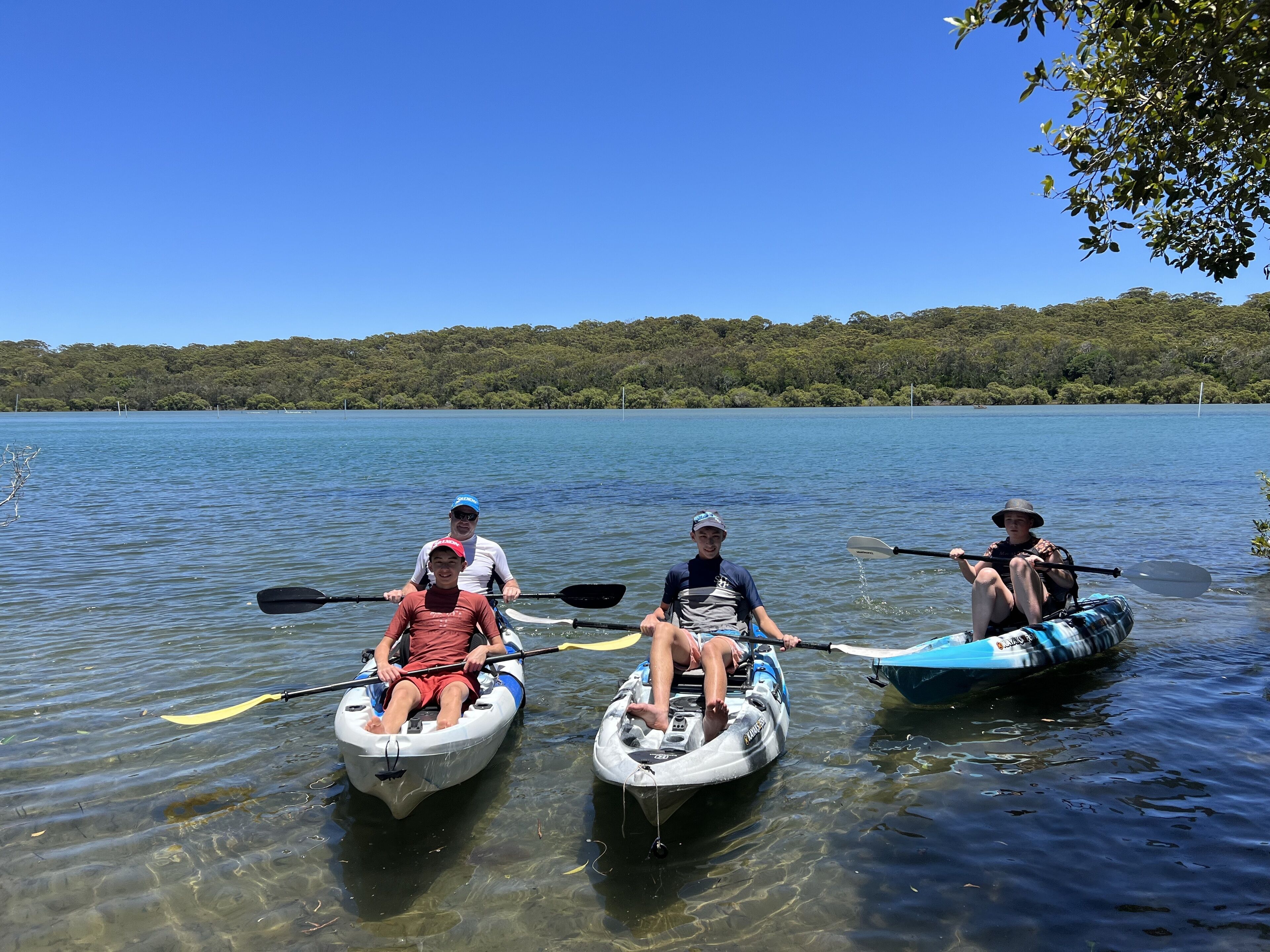 Three people in kayaks on a calm lake with a forested shoreline.