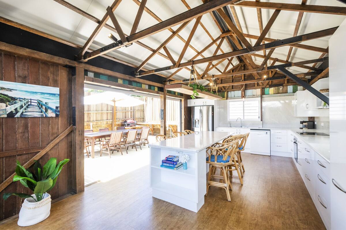 A spacious kitchen with wooden beams, a dining area with wicker chairs, and a view of the ocean through large windows.