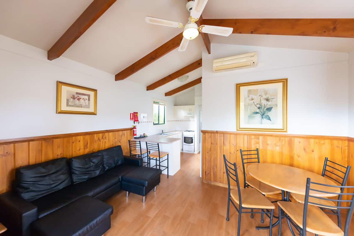 Uma sala de estar espaçosa com um sofá de couro preto, mesa de jantar de madeira e cadeiras. É visível um ventilador de teto e há quadros nas paredes.