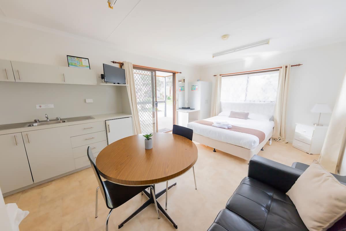 Quarto de hotel moderno com cozinha compacta, área de refeições com mesa e cadeiras de madeira, sofá de couro preto, cama com roupa de cama branca e janelas amplas com cortinas.
