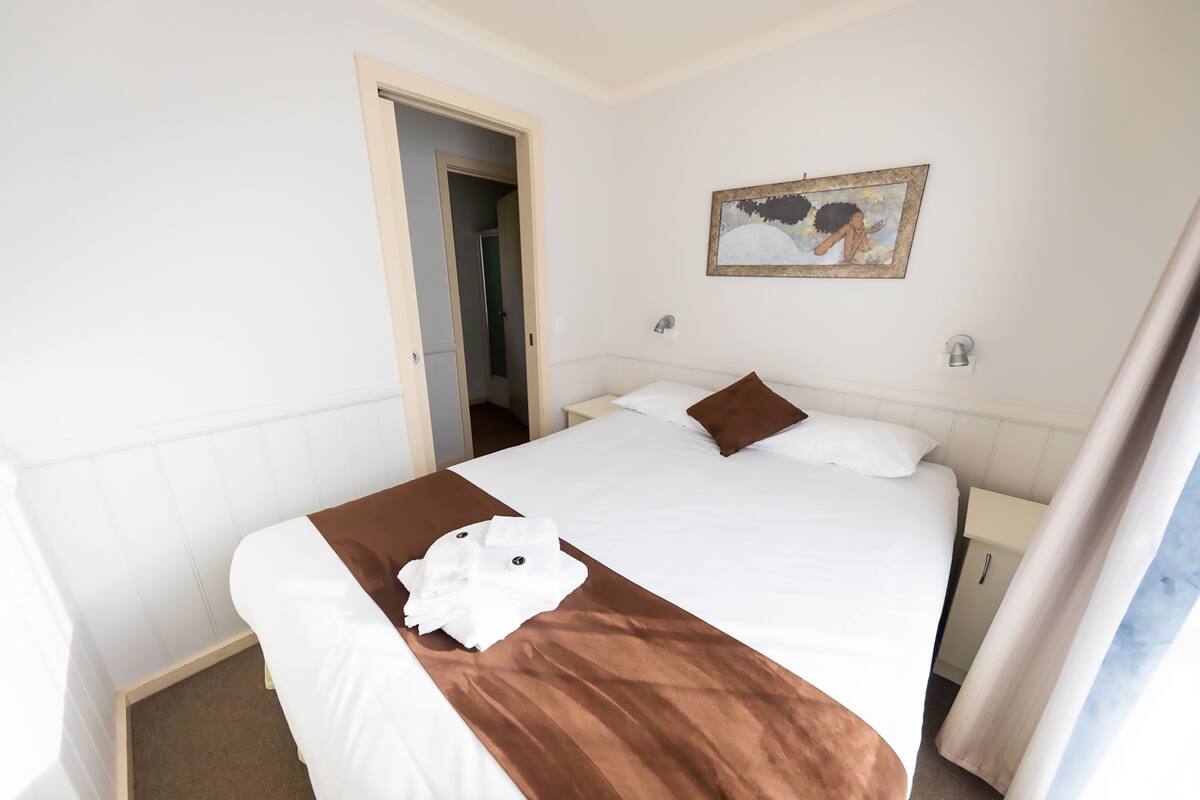 Quarto de hotel com uma cama, um quadro na parede e uma porta que dá para outro quarto.