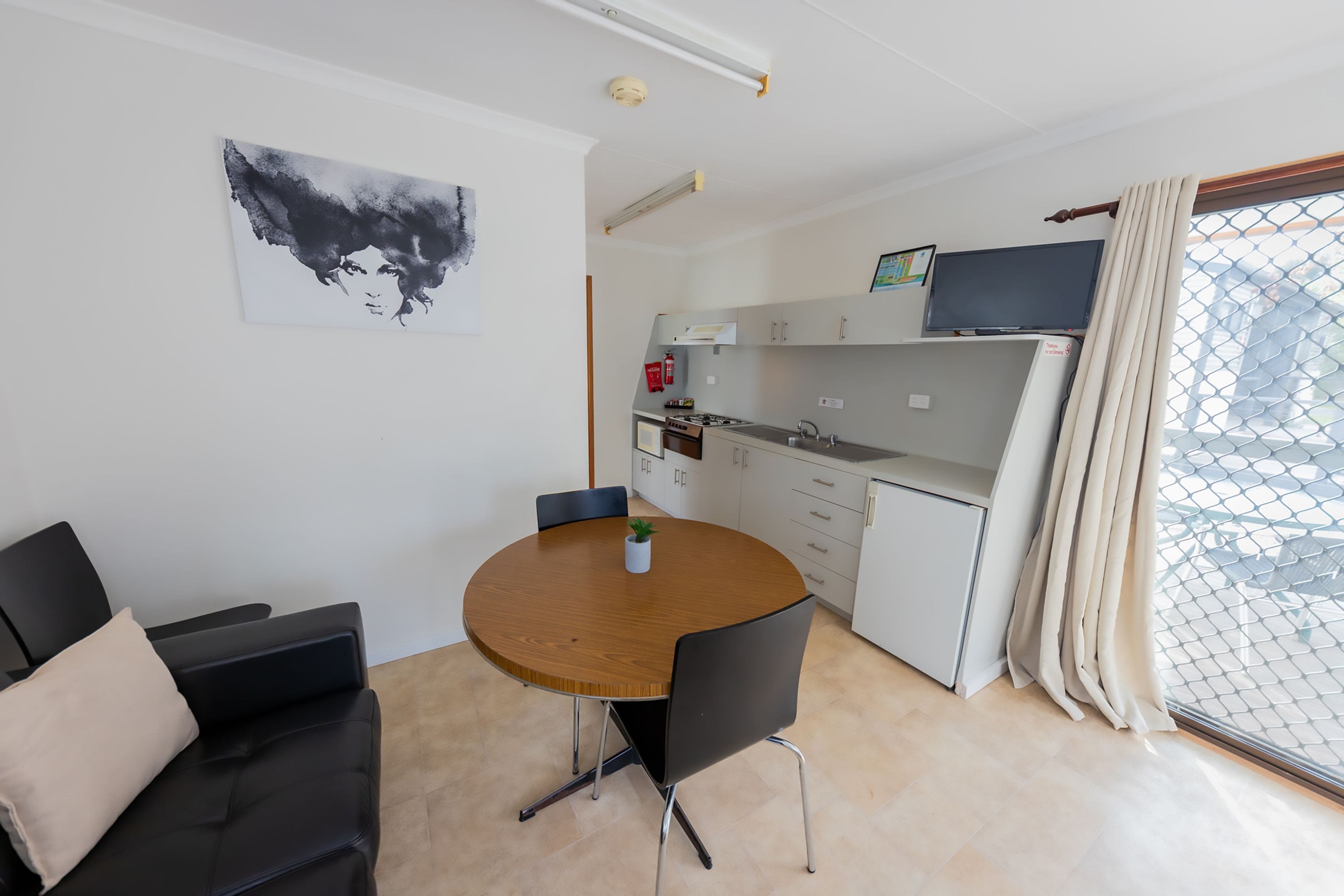 A compact living space with a round wooden dining table, a black sofa, a small potted plant, a kitchenette, and a TV on the wall.
