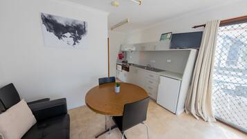 A compact living space with a round wooden dining table, a black sofa, a small potted plant, a kitchenette, and a TV on the wall.