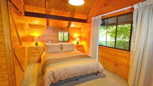 Ein Schlafzimmer einer Holzhütte mit einem Bett, zwei Nachttischlampen, einem Fenster mit Vorhängen und Blick auf Grünfläche.