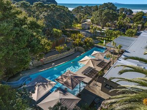 A pool area with umbrellas and lounge chairs, surrounded by trees and a view of the ocean. - Tasman Holiday Parks – Waihi Beach (Waihi Beach)