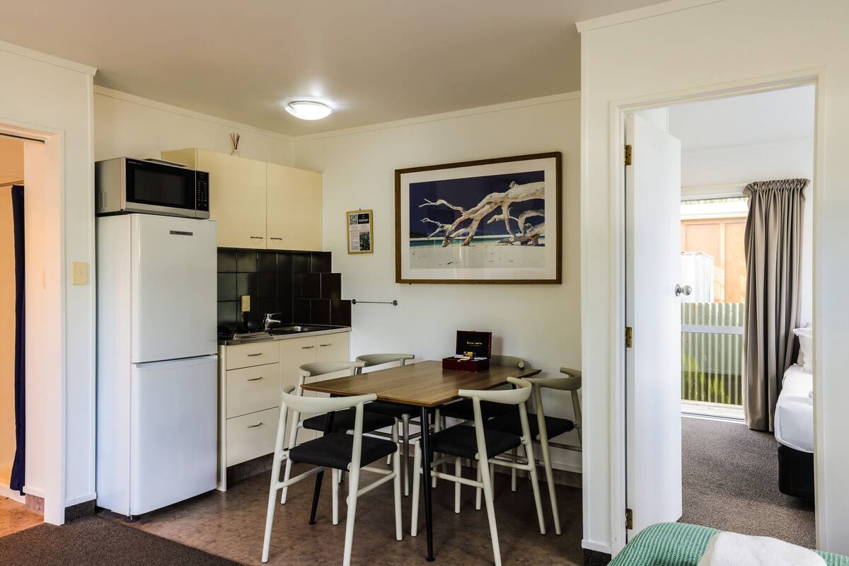 Cocina compacta con microondas, refrigerador y mesa de comedor con sillas. Un cuadro enmarcado cuelga de la pared.
