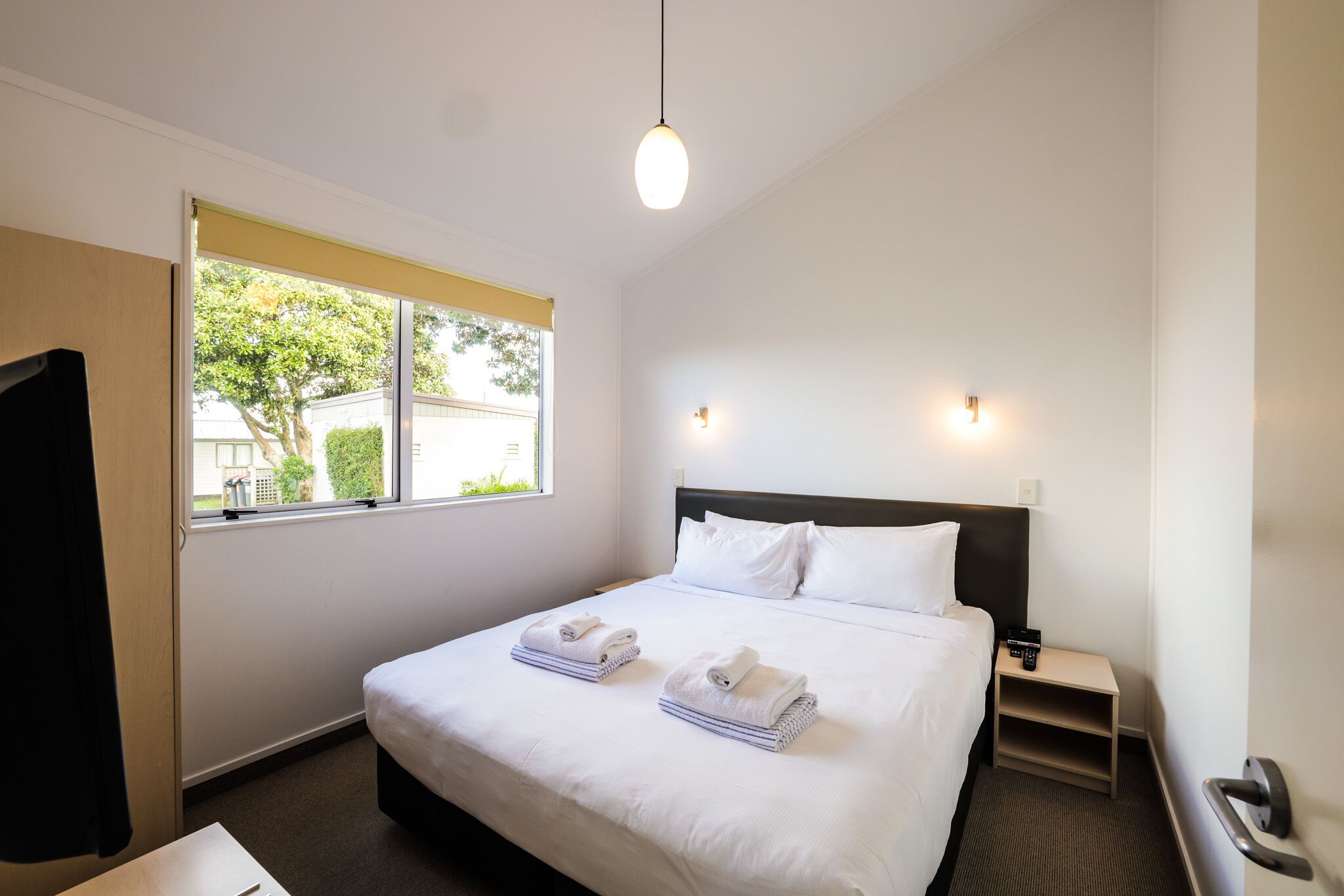 A hotel room with a large bed, two bedside tables, a window with a view of greenery, and a hanging light fixture.