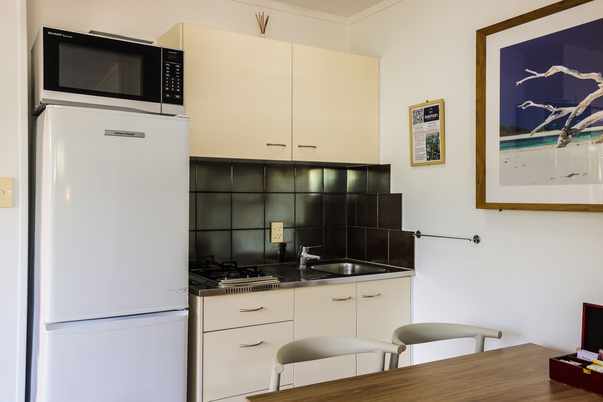 Cocina compacta con refrigerador, microondas, estufa y pileta. Se observa una obra de arte enmarcada en la pared y una mesa pequeña con sillas.