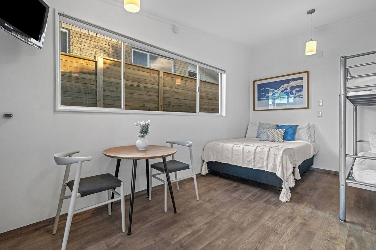 Una habitación pequeña y moderna con una cama, una mesa con dos sillas, un televisor montado en la pared y un ventanal con persianas de madera.