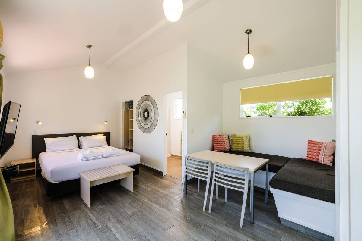 Un dormitorio moderno con cama, mesita de noche y un banco. Hay un comedor con mesa y sillas. Se ve una ventana con vista a zonas verdes.