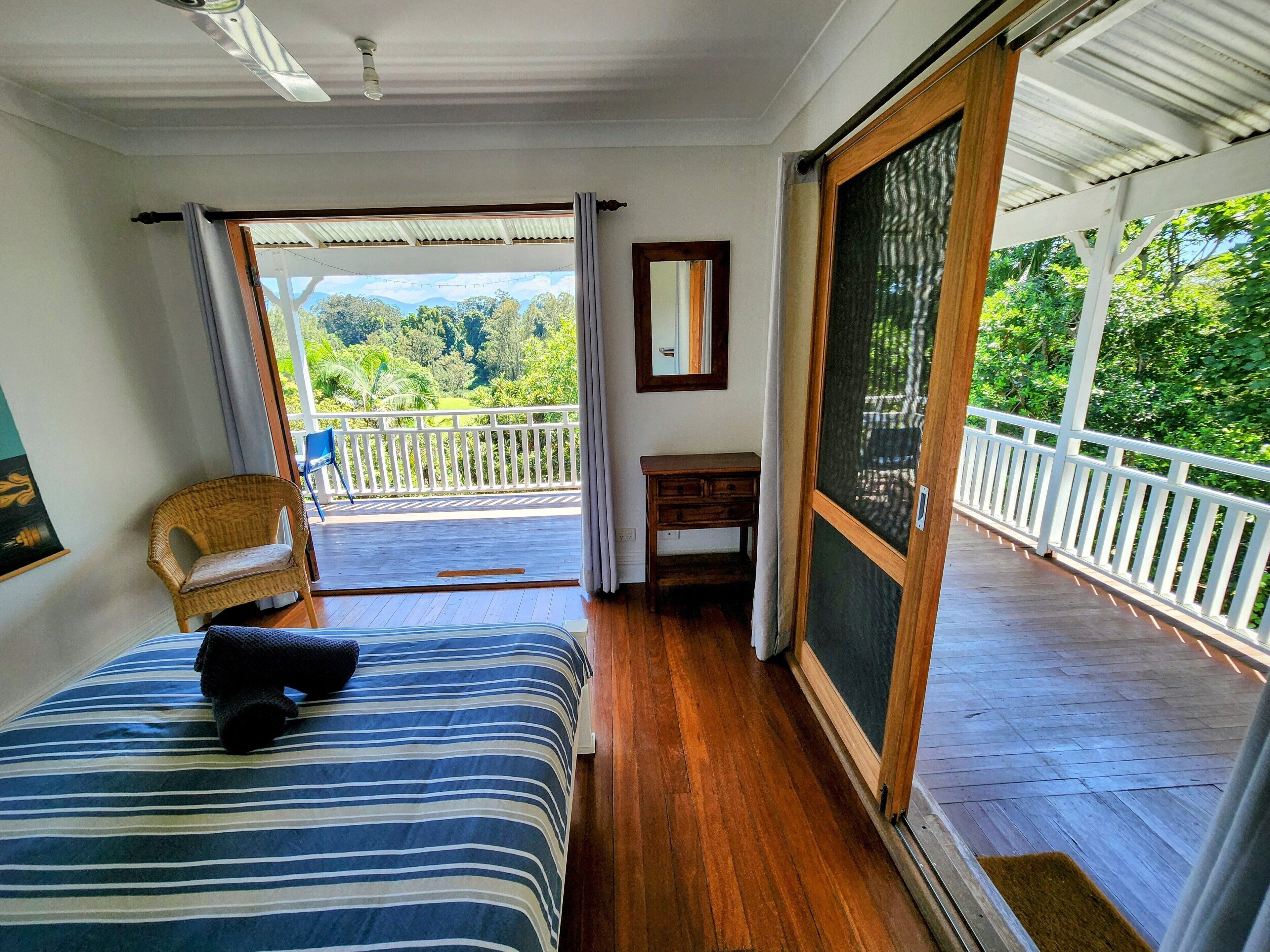 A bedroom with a large bed, a chair, a small table, and a sliding glass door leading to a balcony with a view of greenery.
