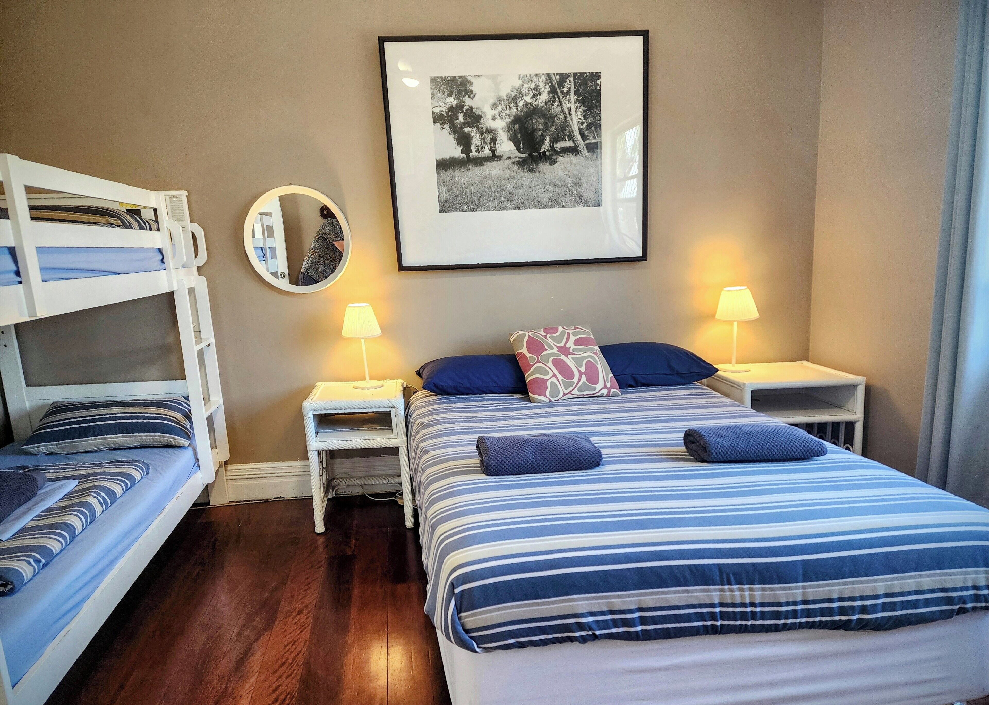 Una habitación con literas, una mesita de noche con lámpara, un cuadro enmarcado en la pared y una ventana con cortinas.