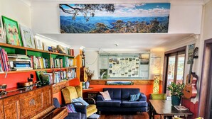 A cozy living room with a piano, bookshelves, a large painting, and a view of a mountainous landscape. - The Belfry Guesthouse (Bellingen)
