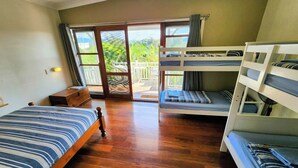 A room with a wooden floor, a single bed with striped bedding, a bunk bed with blue bedding, and a sliding glass door leading to an outdoor area. - The Belfry Guesthouse (Bellingen)