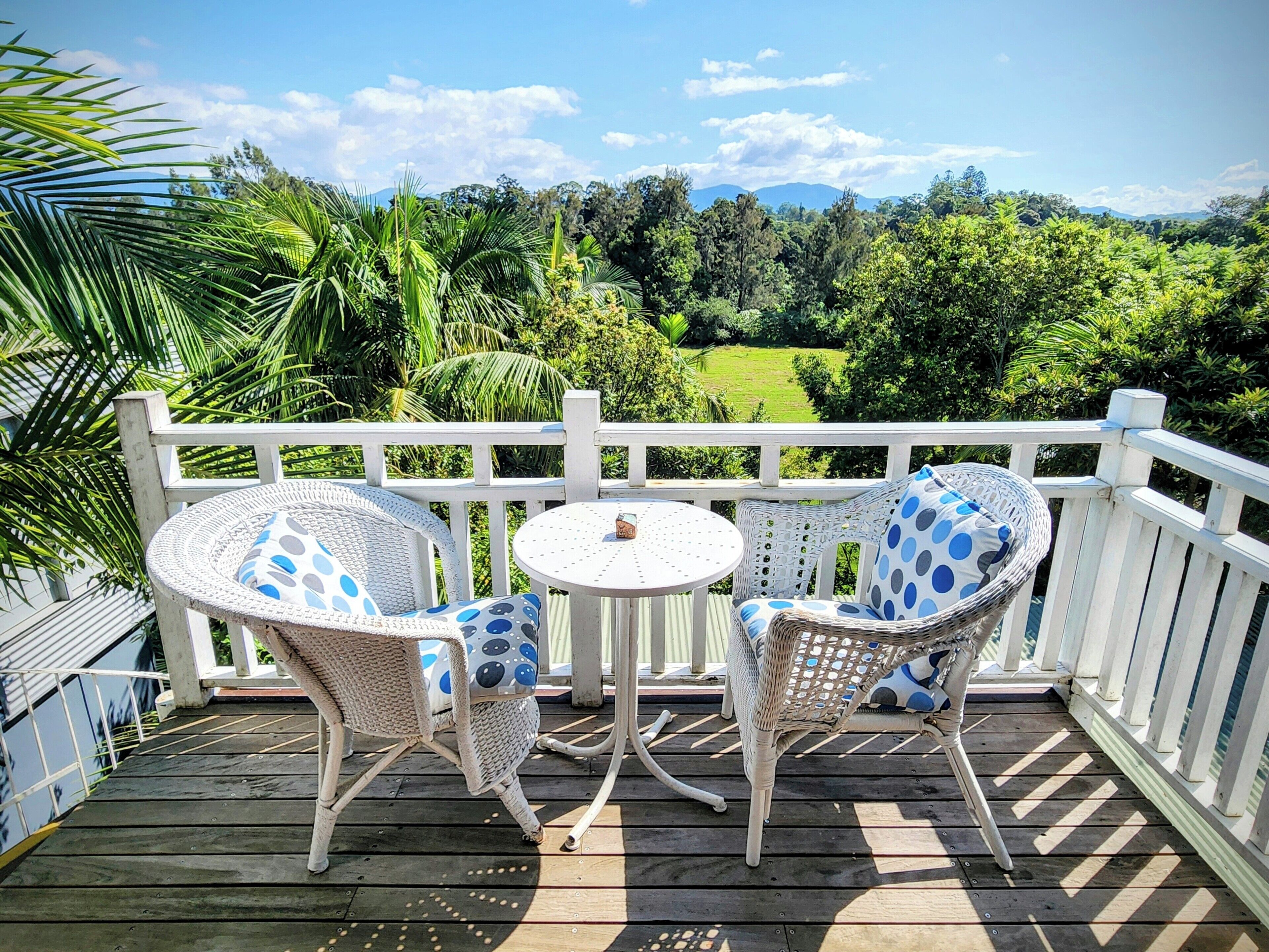 Une terrasse en bois avec deux fauteuils en osier et une petite table ronde, offrant une vue sur un paysage verdoyant et luxuriant, avec des arbres et une chaîne de montagnes au loin.